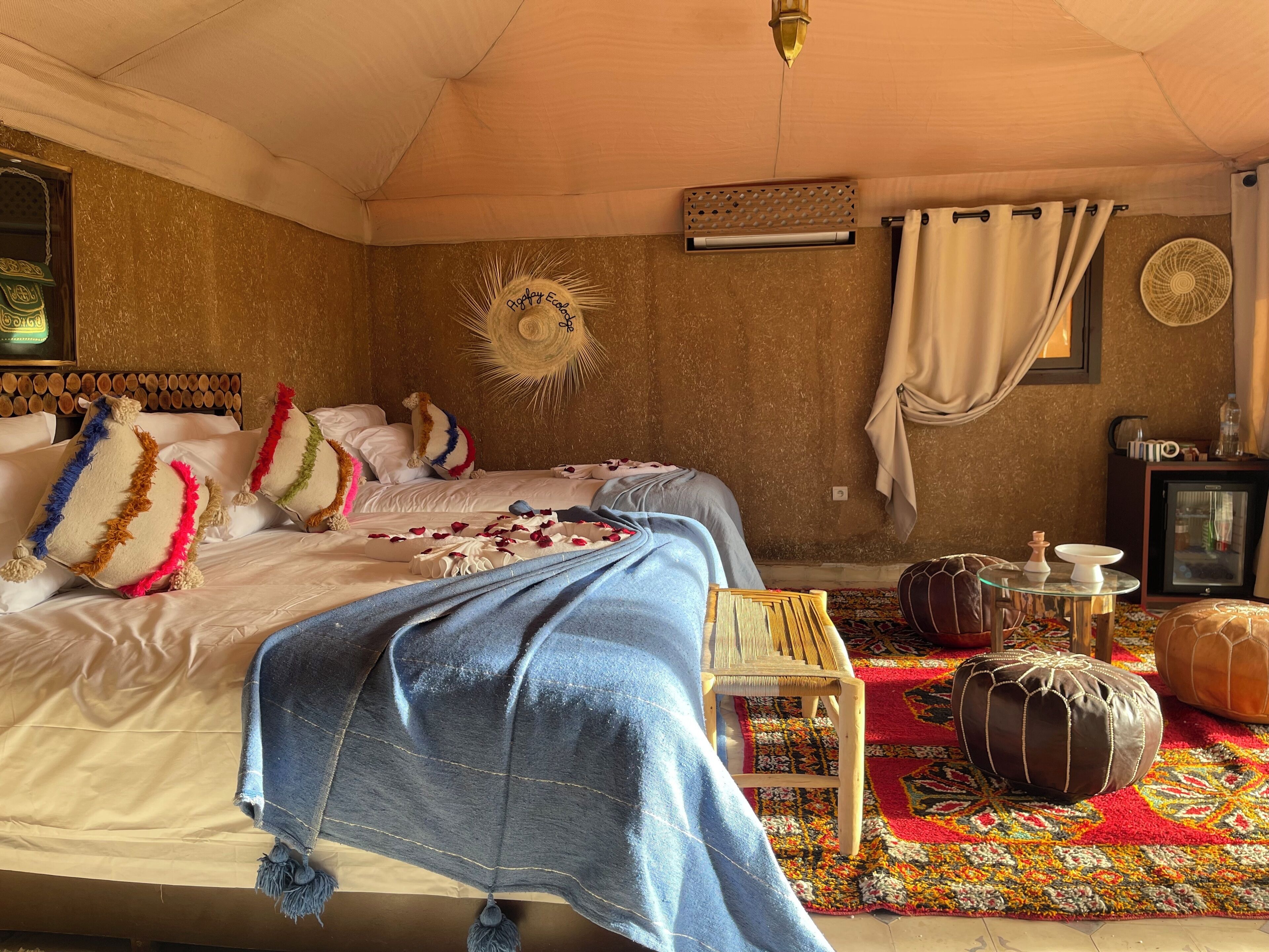Een knus slaapkamer met een bed, kussens, een zonvormige decoratie aan de muur, een tafeltje met een ventilator en een kleurrijk vloerkleed.