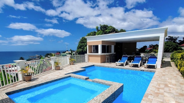 A swimming pool with a view of the ocean, a covered seating area with chairs, and a white fence. - Stunning Ocean, City & Pool View 2 Bedroom Apt. 2 minutes from Roseau (Roseau)