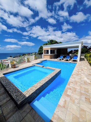 A swimming pool with a view of the ocean, a covered seating area with chairs, and a white fence. - Stunning Ocean, City & Pool View 2 Bedroom Apt. 2 minutes from Roseau (Roseau)