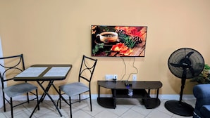 A room with a dining table, two chairs, a flat-screen TV mounted on the wall, a small table with a fan, and a blue sofa. - Stunning Ocean, City & Pool View 2 Bedroom Apt. 2 minutes from Roseau (Roseau)