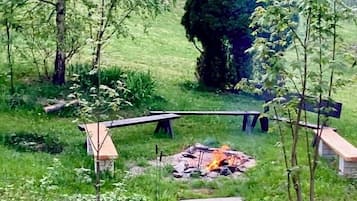 Ein grünes Außenareal mit Feuerstelle, Bänken und einer Steintreppe, die zu einer Holterrasse führt.