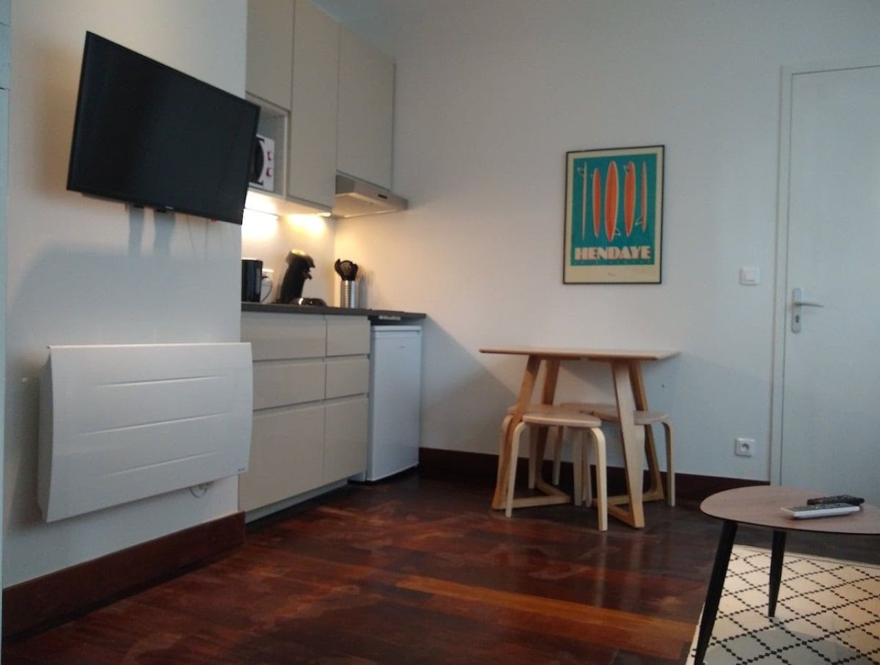 Une cuisine compacte équipée d’un lave-vaisselle intégré, une petite table à manger avec des chaises, un téléviseur fixé au mur et une affiche accrochée au mur.