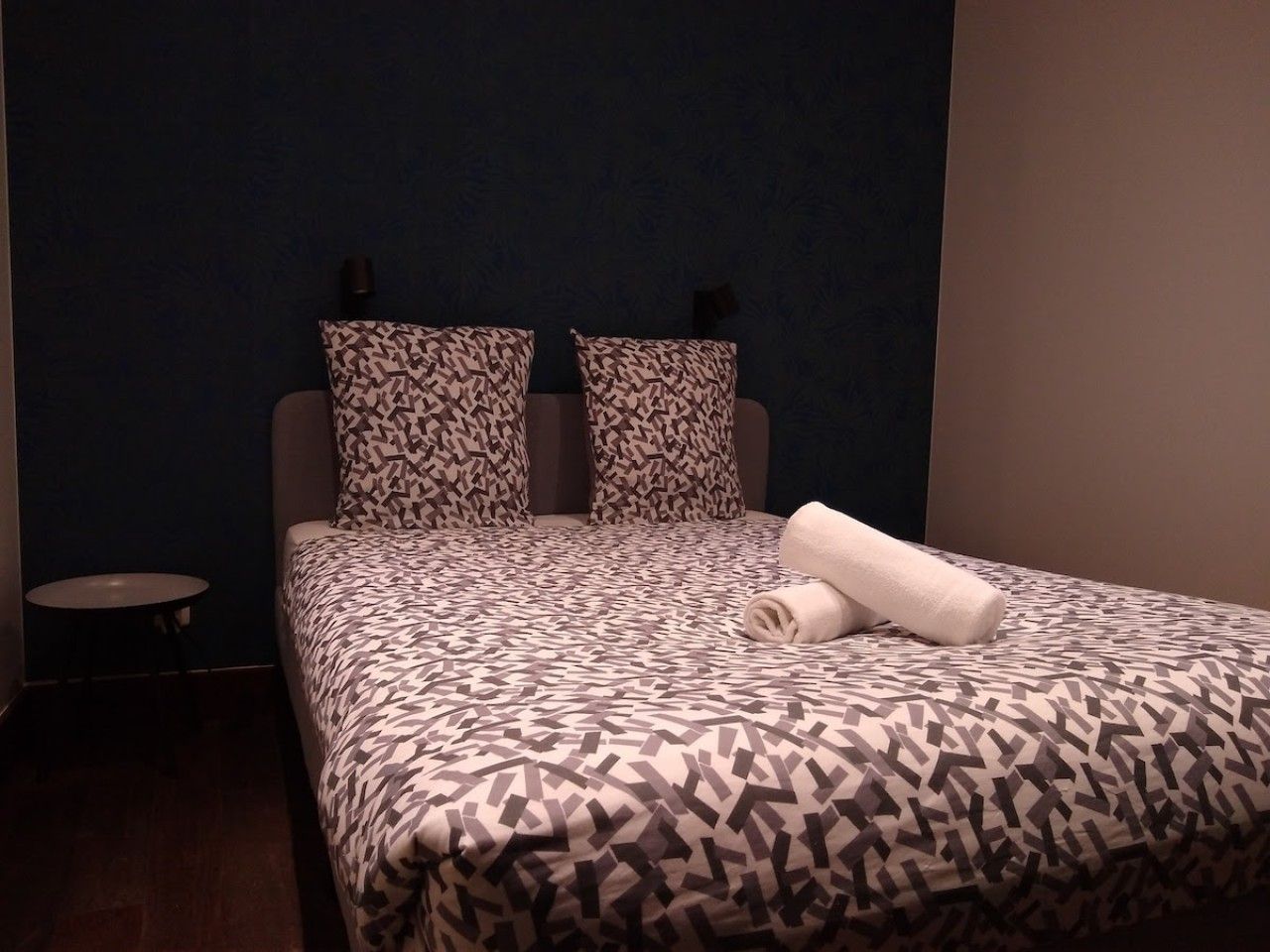 Un lit bien fait, avec une literie à motifs, une petite table ronde et un mur orné d’un motif bleu foncé.