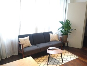 A living room with a sofa, a small table, a potted plant, and a window with curtains. - Two Bedroom apartment in historical  center (Bordeaux)