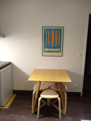 A small room with a wooden table and two chairs, a refrigerator, and a wall-mounted poster. - Two Bedroom apartment in historical  center (Bordeaux)