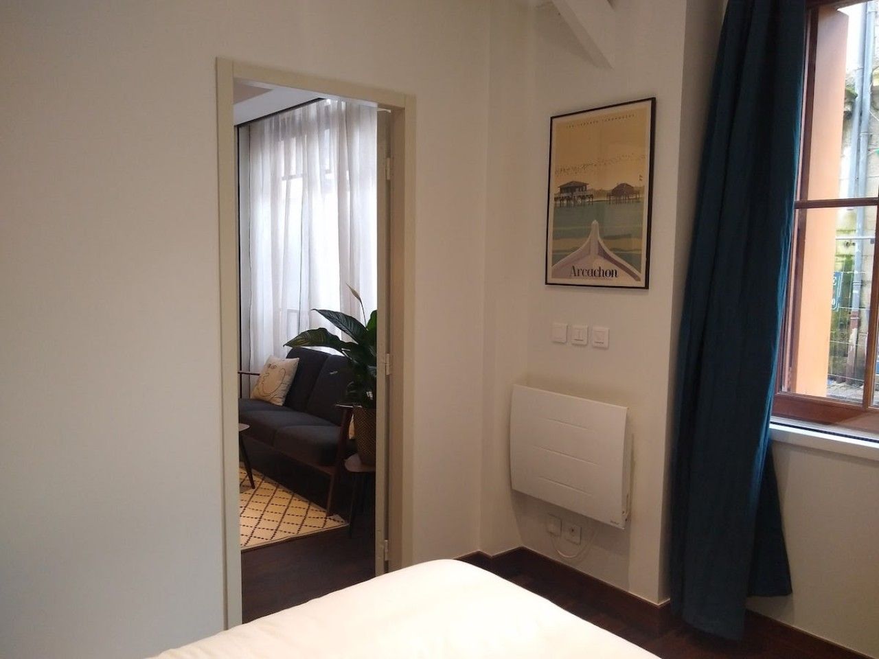 Une chambre d’hôtel avec un lit, un radiateur, une fenêtre avec des rideaux, un tableau encadré au mur et une vue sur une autre pièce où se trouvent un canapé et des rideaux.
