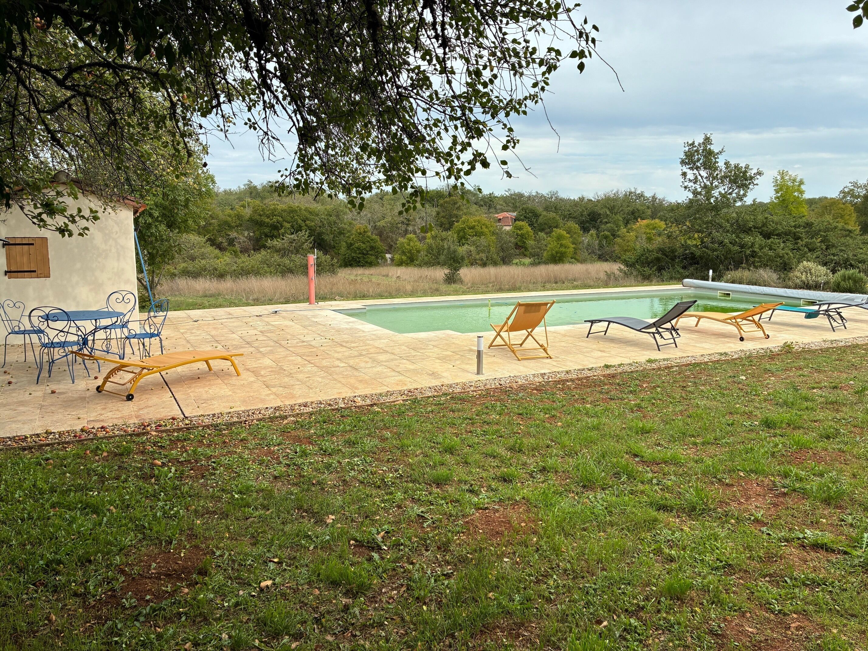 A pool area with lounge chairs and a table, surrounded by greenery and a small building.