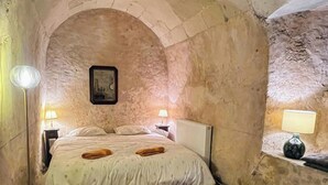Una habitación pequeña y estrecha, con paredes de piedra, una cama, dos lámparas de noche y una mesita con lámpara.