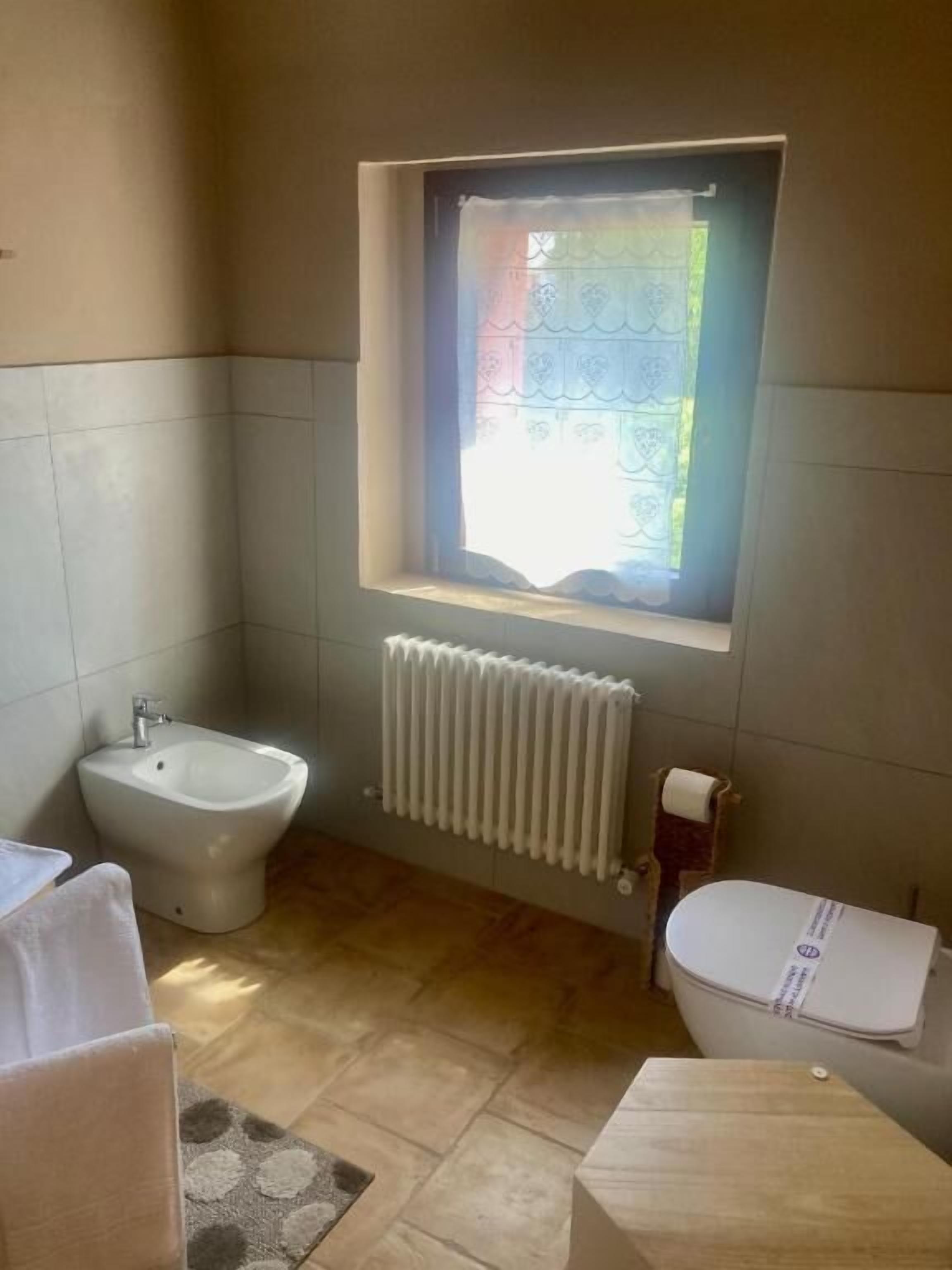Un baño con bidé, inodoro, radiador y ventana con cortina.