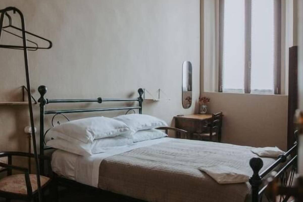 A bedroom with a bed, a chair, a small table, a mirror, and a window with curtains.