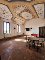 Una habitación espaciosa con techo alto y frescos elaborados, una mesa redonda de madera con sillas, un sofá rojo y un televisor de pantalla plana montado en la pared.