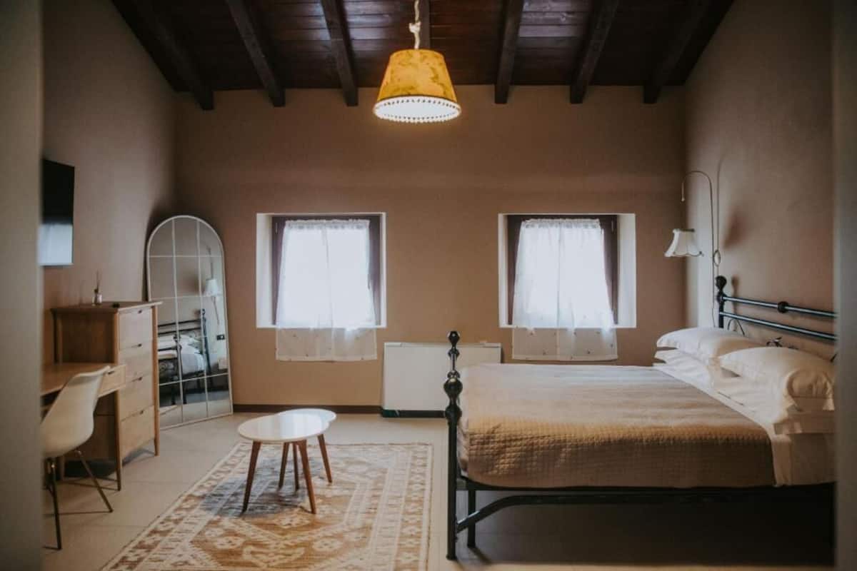 A bedroom with a bed, a desk, two windows, a radiator, and a hanging lamp.