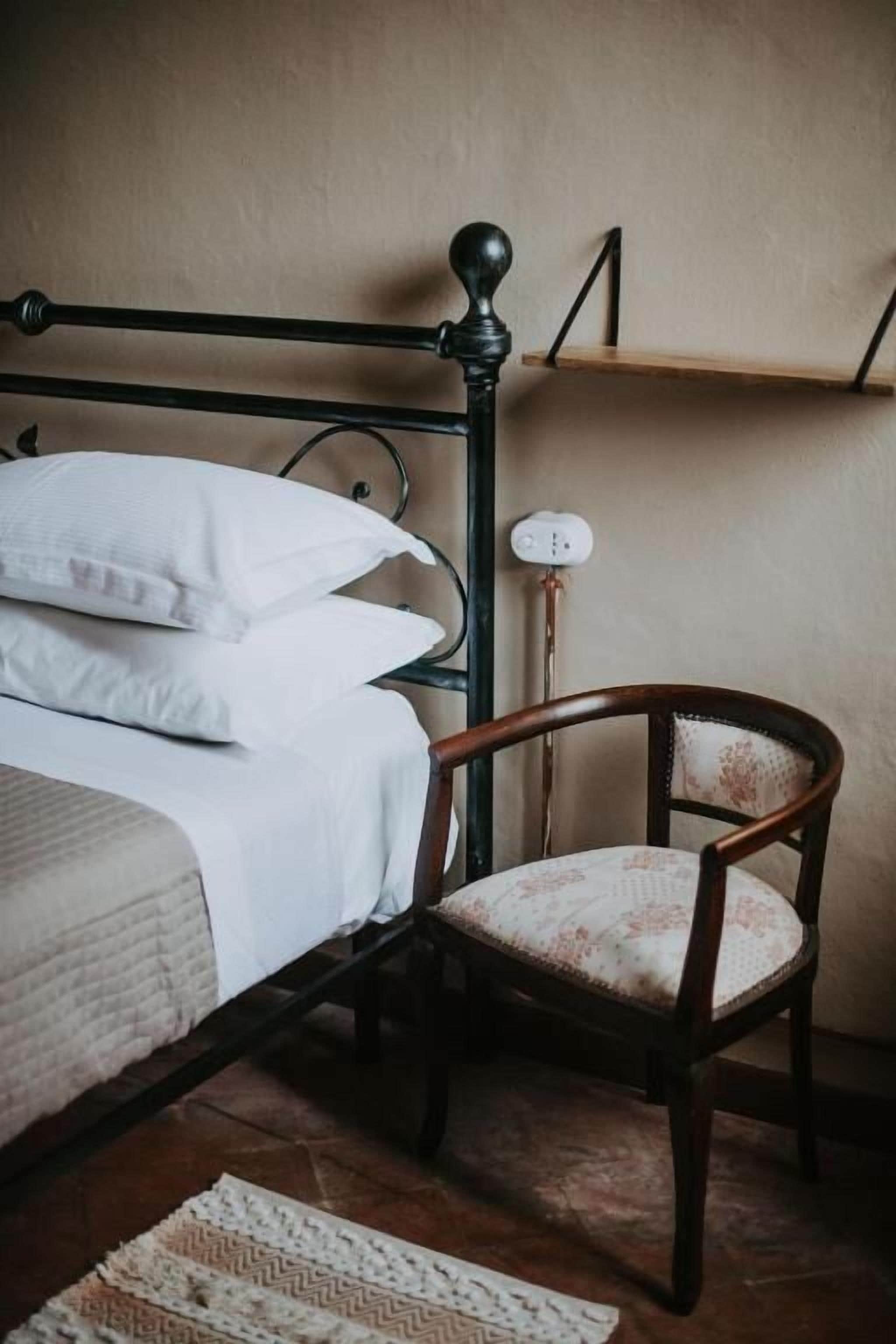 Um quarto com cama, cadeira, mesa de cabeceira e prateleira.