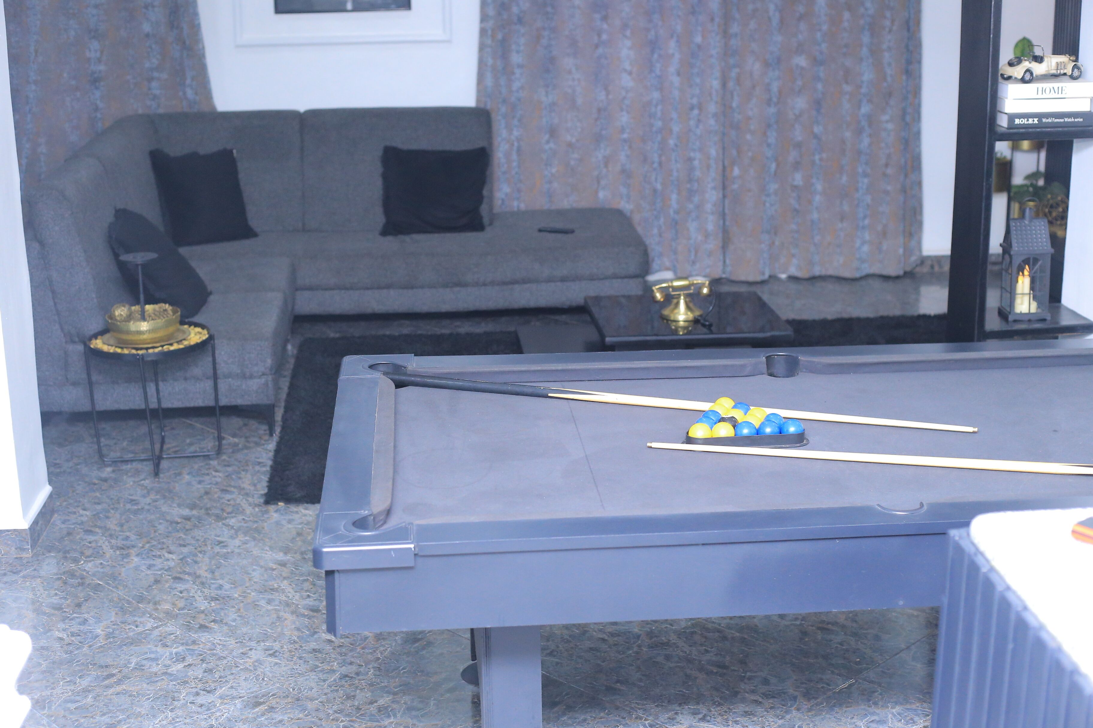 A pool table with billiard balls, a sofa with black cushions, a small side table with a lamp, and a bookshelf with decorative items.