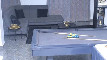 A pool table with billiard balls, a sofa with black cushions, a small side table with a lamp, and a bookshelf with decorative items.