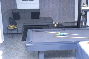 A pool table with billiard balls, a sofa with black cushions, a small side table with a lamp, and a bookshelf with decorative items.