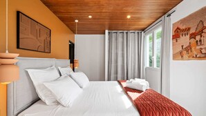 Een slaapkamer met een houten plafond, een bed met witte beddengoed, een rode geborduurde plaid en een raam met gordijnen.