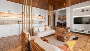 Een moderne woonkamer met een bank, salontafel en houten details. Achtergrond toont een keuken met witte kasten en een aanrecht.