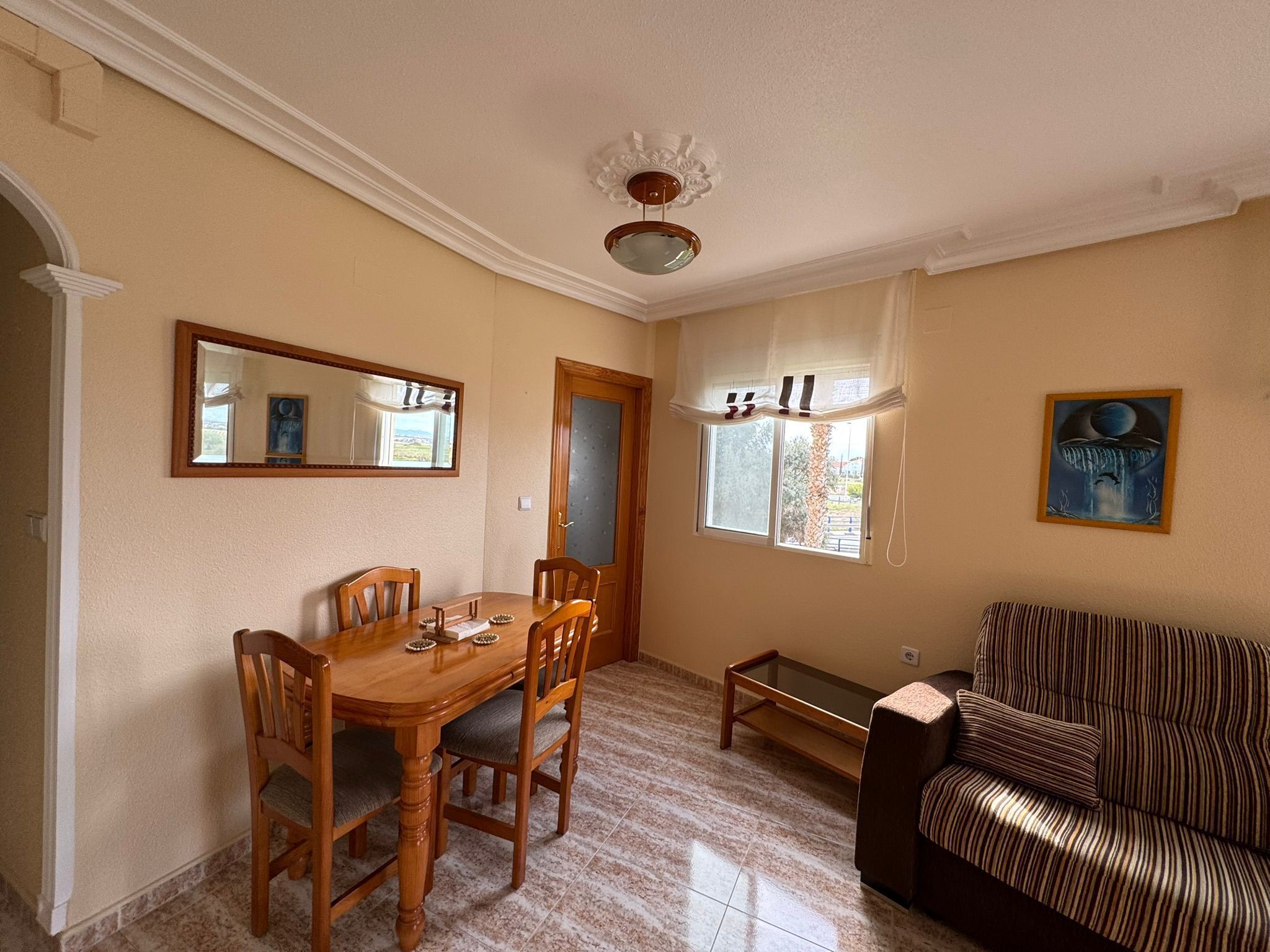A dining area with a wooden table and chairs, a small side table, a sofa, a window with curtains, and a framed picture on the wall.