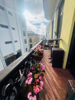 Ein Balkon mit Blumentöpfen und Korbmöbeln, mit Blick auf eine Straße mit Gebäuden.