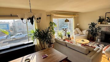 A cozy living room with a sofa, a coffee table, and a dining table. There are plants and a fireplace.