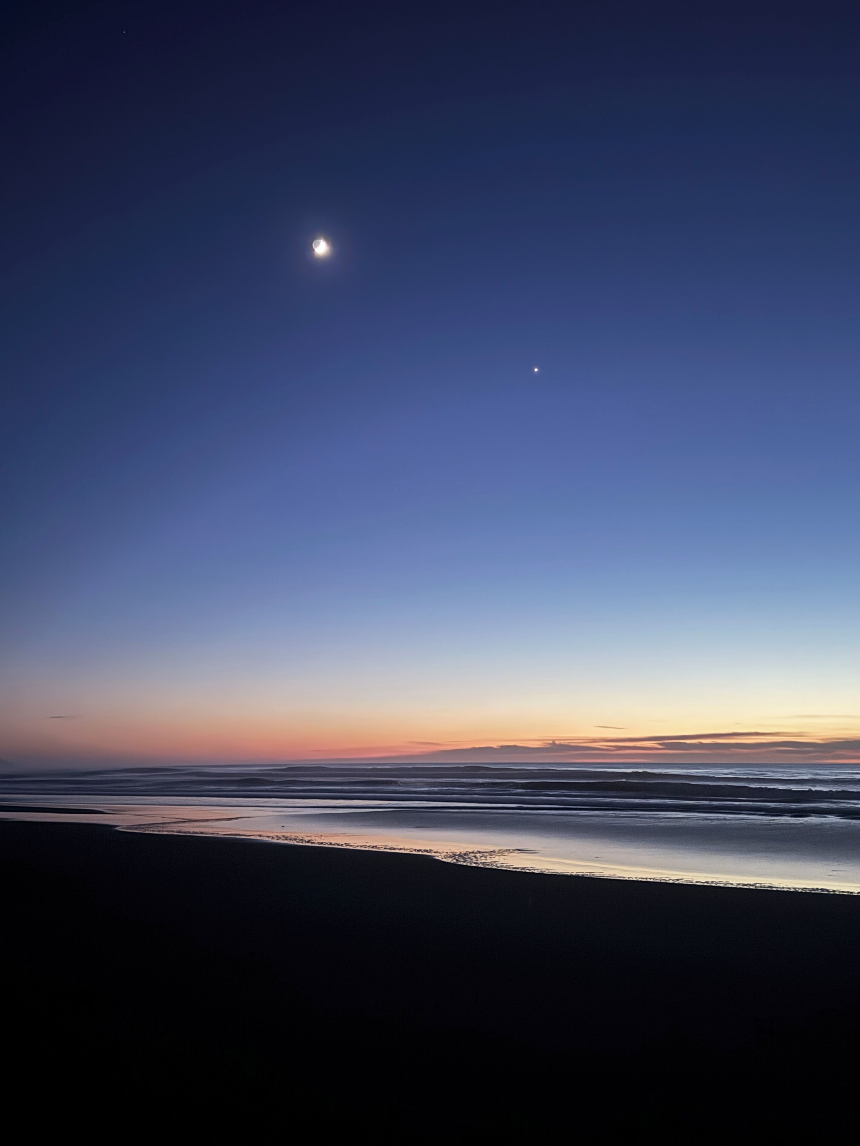 A beach at dusk with a clear sky, calm sea, and a visible moon.