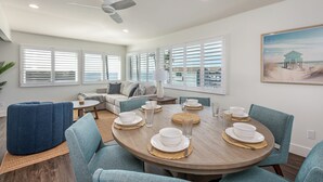 Ein Essbereich mit einem runden Tisch, gedeckt für sechs Personen, umgeben von blauen Stühlen und Blick auf den Strand durch große Fenster.