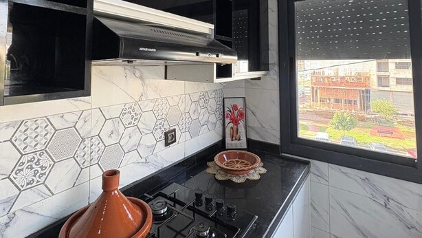 A compact kitchen with a gas stove, black cabinets, a tiled backsplash, and a window with a view of the outside. - Gulf Serenity Suites (Fès)