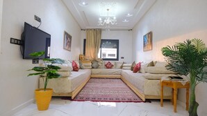 A living room with a sectional sofa, a flat-screen TV, a chandelier, and a large potted plant. - Gulf Serenity Suites (Fès)