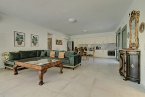 A living room with a green sofa, a wooden coffee table, and a dining area with a white table and chairs. There is a kitchen with white cabinets and a black and gold cabinet.
