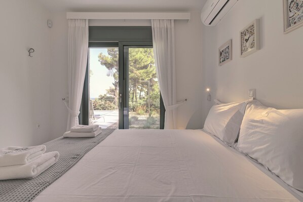 A bedroom with a bed, white curtains, a view of greenery outside, and a wall-mounted air conditioner.
