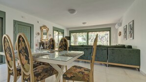 Una zona de menjador amb una taula de vidre, cadires tapissades amb motius florals, un sofà verd i vista cap a l'exterior a través d'una finestra gran.