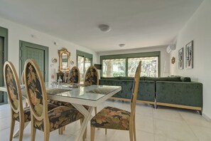 A dining area with a glass-top table, floral upholstered chairs, a green sofa, and a view of the outdoors through a large window. - Ouzo Stone House2 with Private Pool and Parking (Plomari)