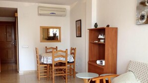 Een woonkamer met een eettafel, stoelen, een bank, een klein bijzettafeltje, een houten boekenkast en een airconditioner.