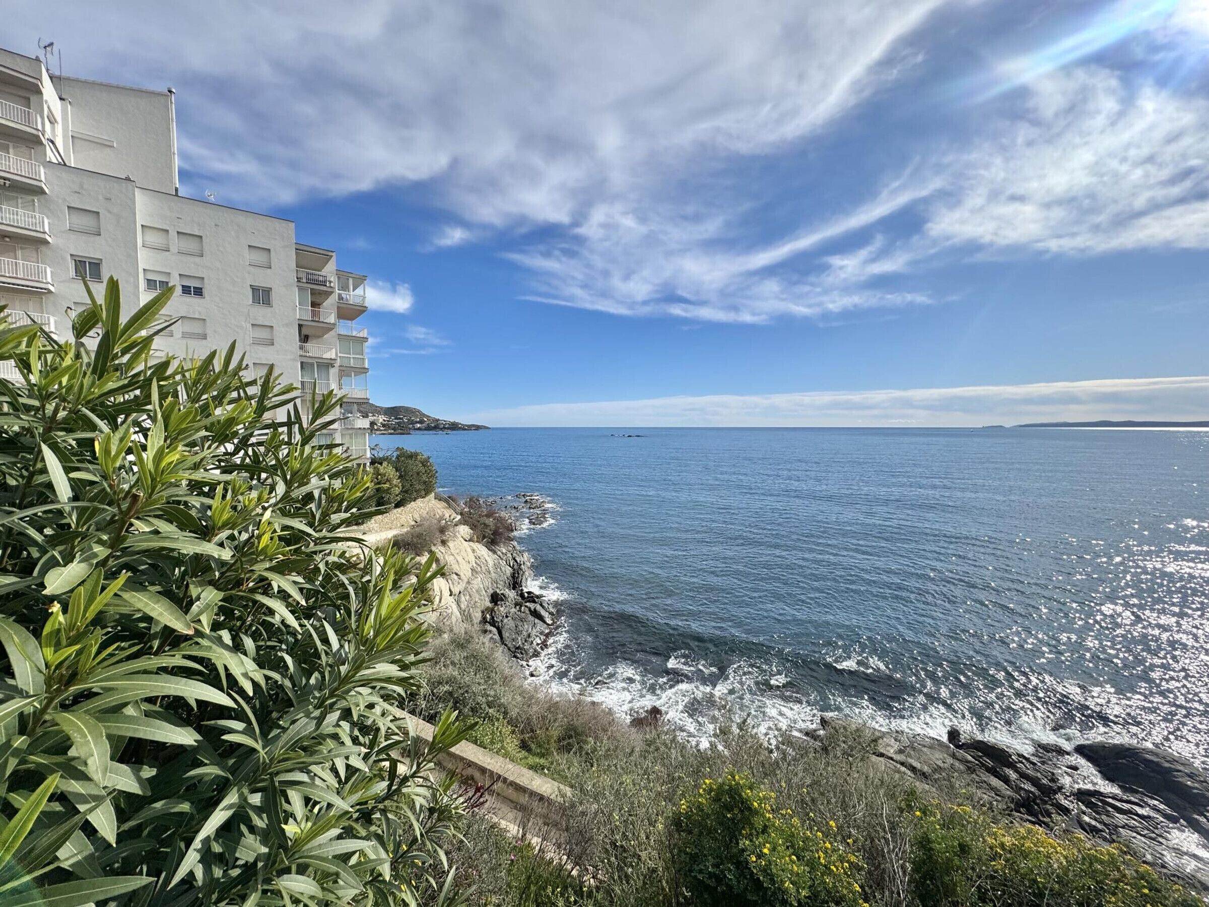 A coastal view with a building adjacent to the sea, featuring balconies and a clear sky.