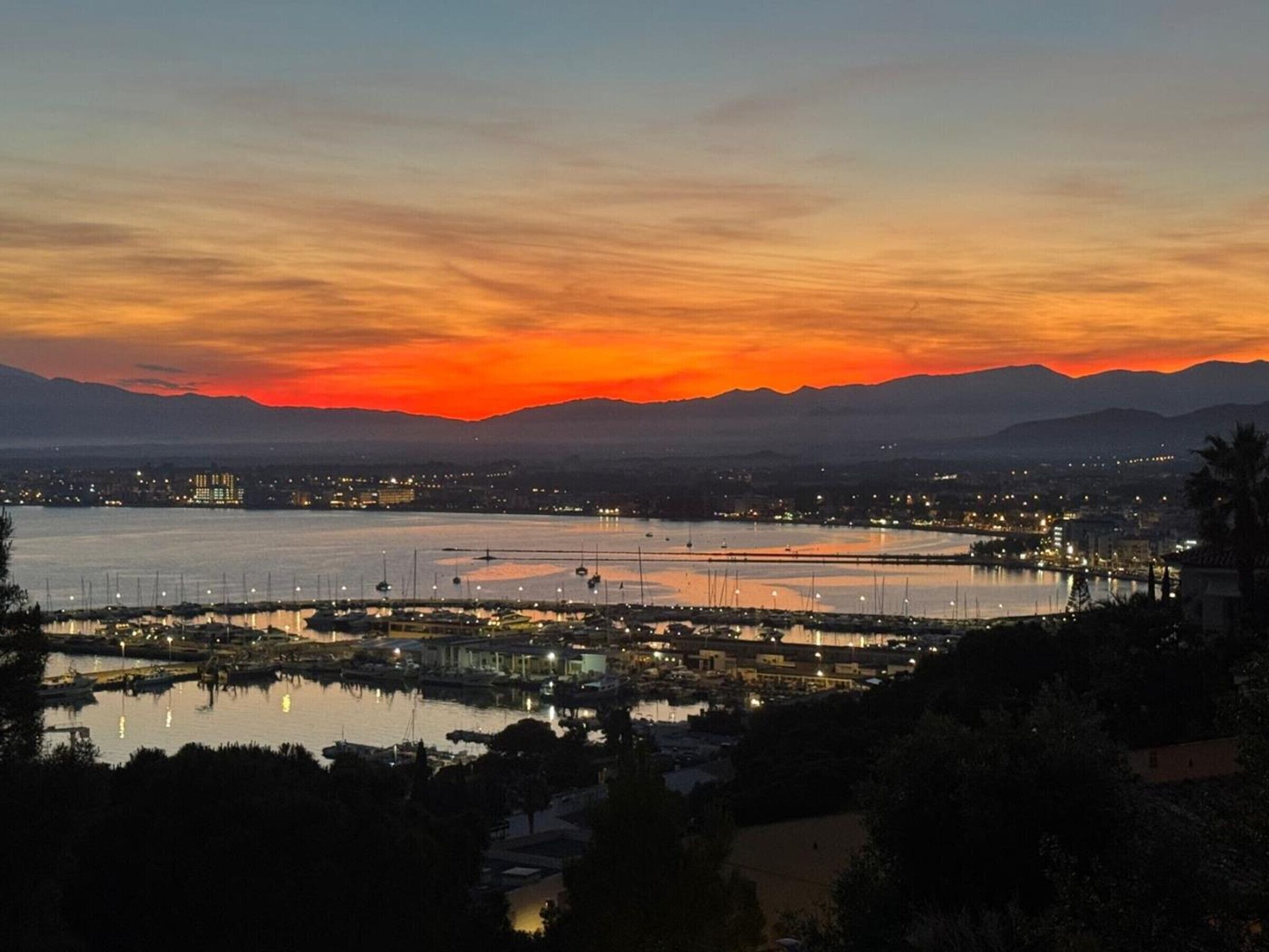 A coastal cityscape at dusk with a sunset over mountains, a marina with boats, and distant city lights.