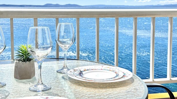 A table with a glass top, a floral plate, and two wine glasses, set against a backdrop of a clear blue sea and distant mountains. - Beautiful apartment with pool, parking, and views of the Bay of Roses (Roses)