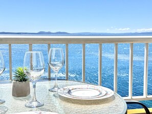 A table with a glass top, a floral plate, and two wine glasses, set against a backdrop of a clear blue sea and distant mountains. - Beautiful apartment with pool, parking, and views of the Bay of Roses (Roses)
