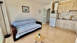 A modern apartment with a sofa, a small round table with a plant, a kitchen with wooden cabinets, and a framed picture on the wall. - Beautiful apartment with pool, parking, and views of the Bay of Roses (Roses)