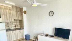 A modern kitchen with wooden cabinets, a white countertop, a ceiling fan, a clock, and a TV on a white cabinet. - Beautiful apartment with pool, parking, and views of the Bay of Roses (Roses)