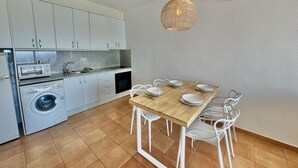 A compact kitchen with a dining area, including a washing machine, microwave, and a hanging light fixture. - Apartment with pool and parking 700m from the beach (Roses)