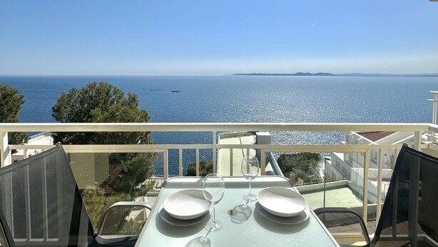 A table with two plates and two wine glasses on a balcony overlooking a clear blue sea with distant land and a few boats. - Apartment with pool and parking 700m from the beach (Roses)