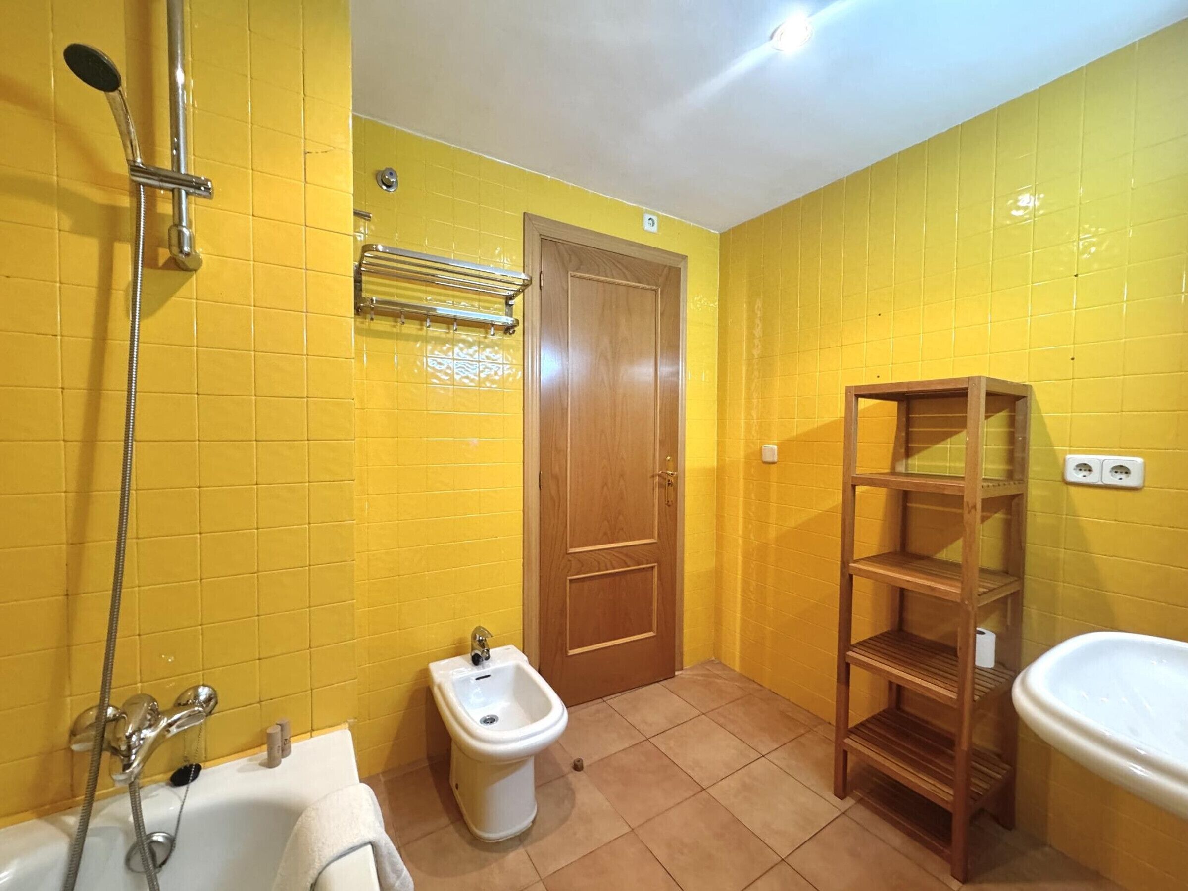 A bathroom with yellow tiled walls, a wooden door, a bidet, a shelf, and a bathtub.