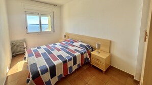 A bedroom with a bed, a nightstand, a lamp, a ceiling fan, a window with a view, and a radiator. - Apartment with pool and parking 700m from the beach (Roses)