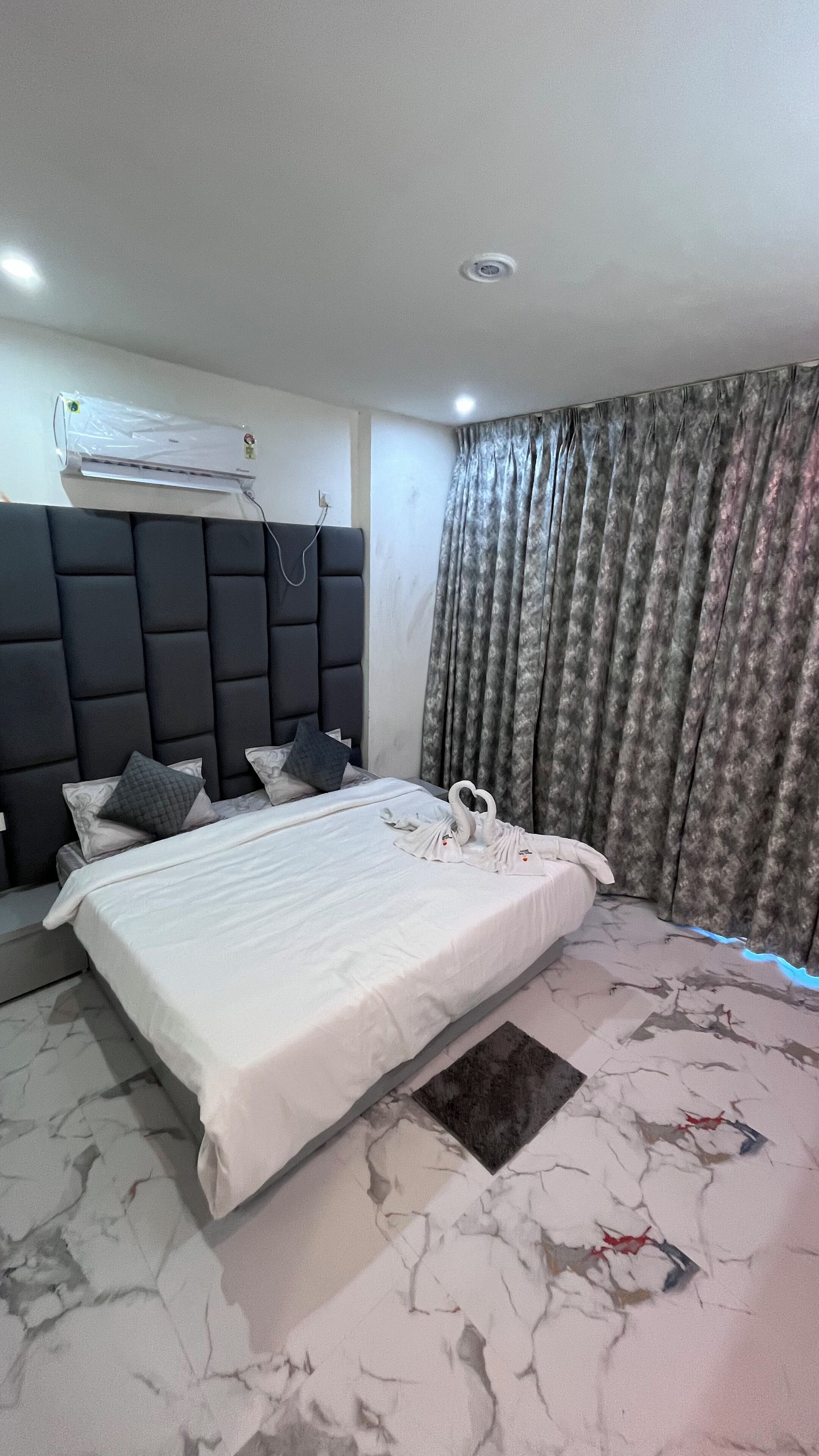 A modern hotel room with a large bed, a headboard, a ceiling-mounted air conditioner, and a window with curtains.