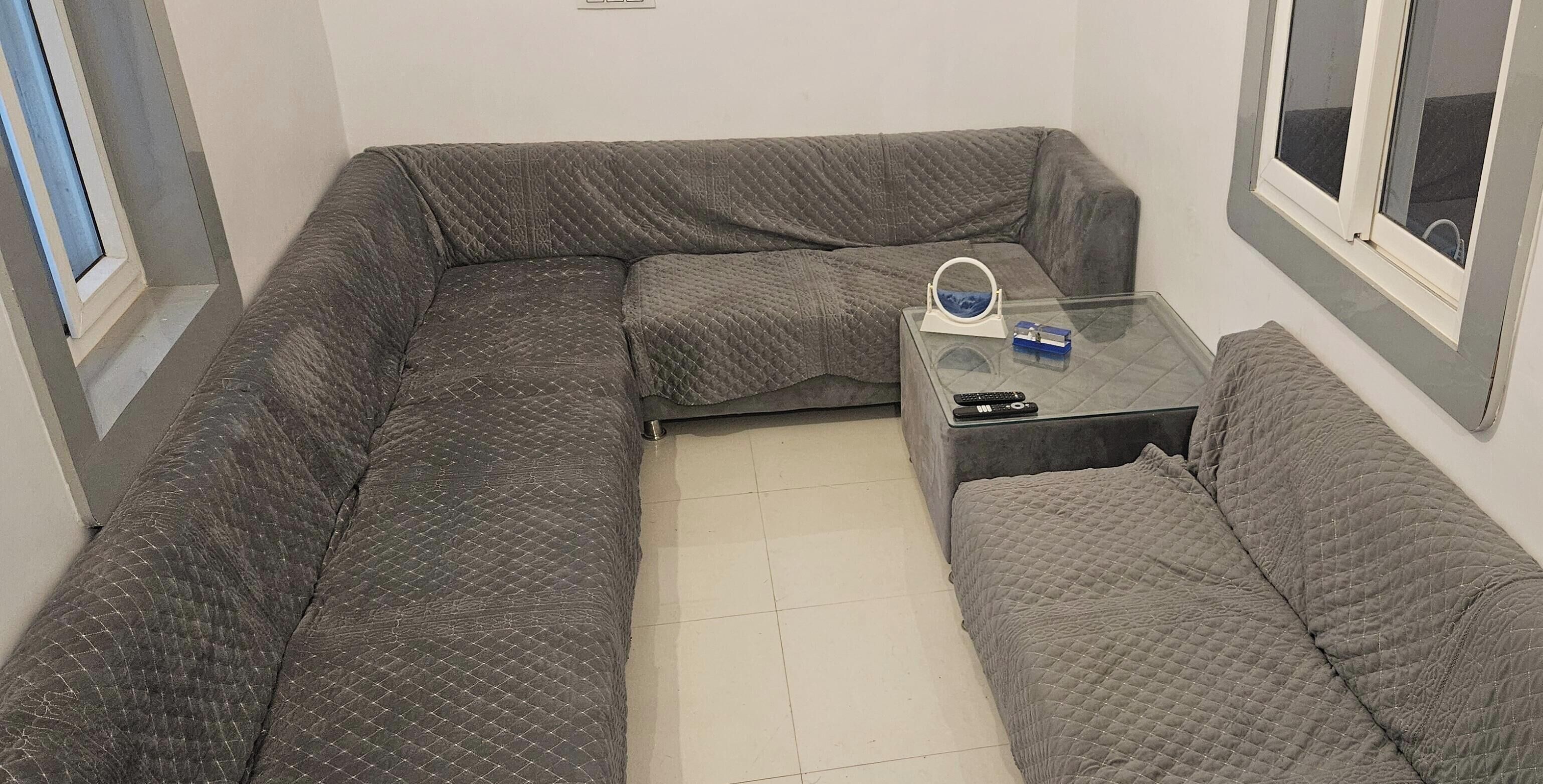 Um espaço de estar compacto com um sofá cinza acolchoado, uma mesa de centro de vidro e um pequeno apoio para copos.