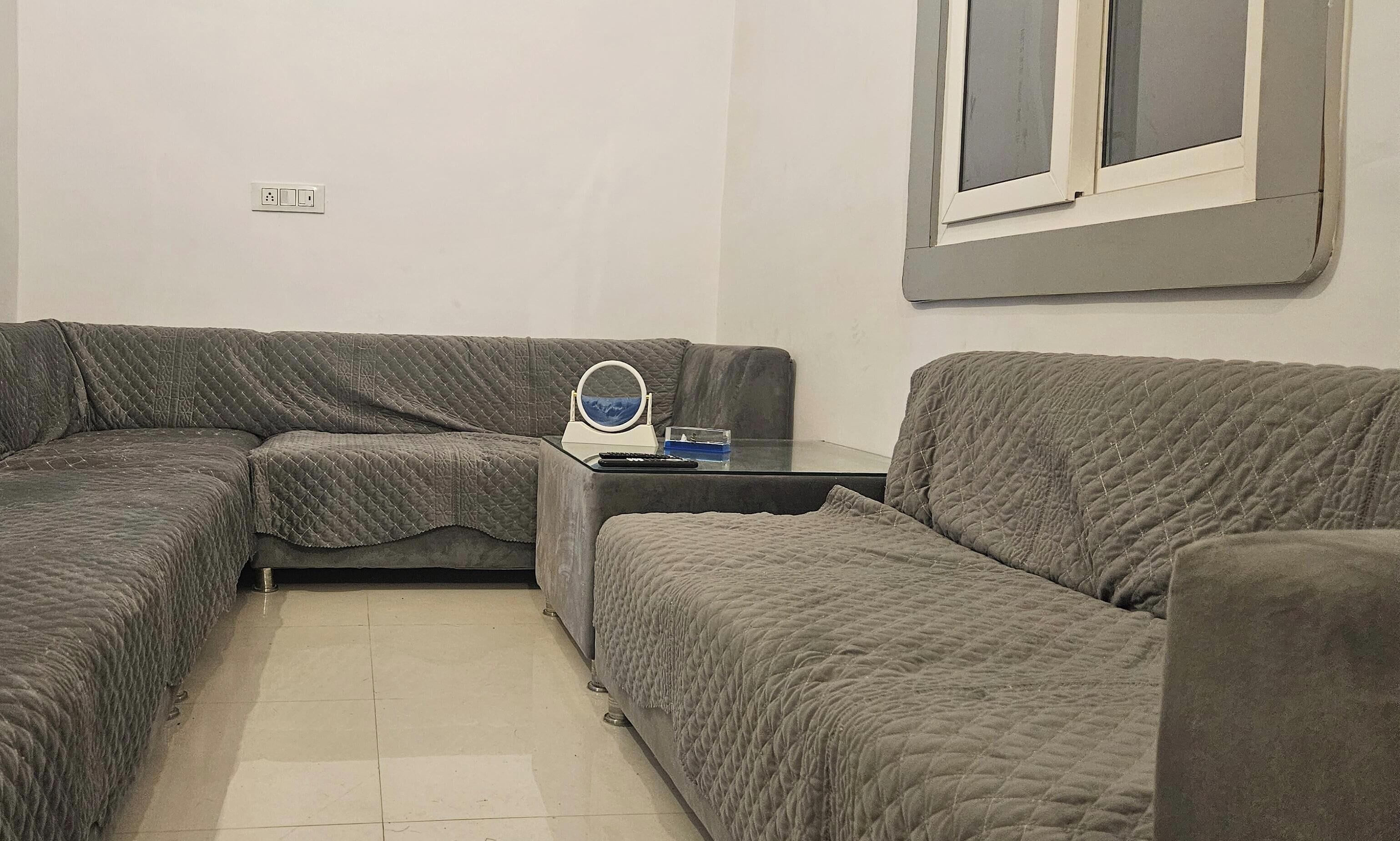 A living room with a grey sectional sofa, a small side table with a mirror and a remote control, and a window with blinds.