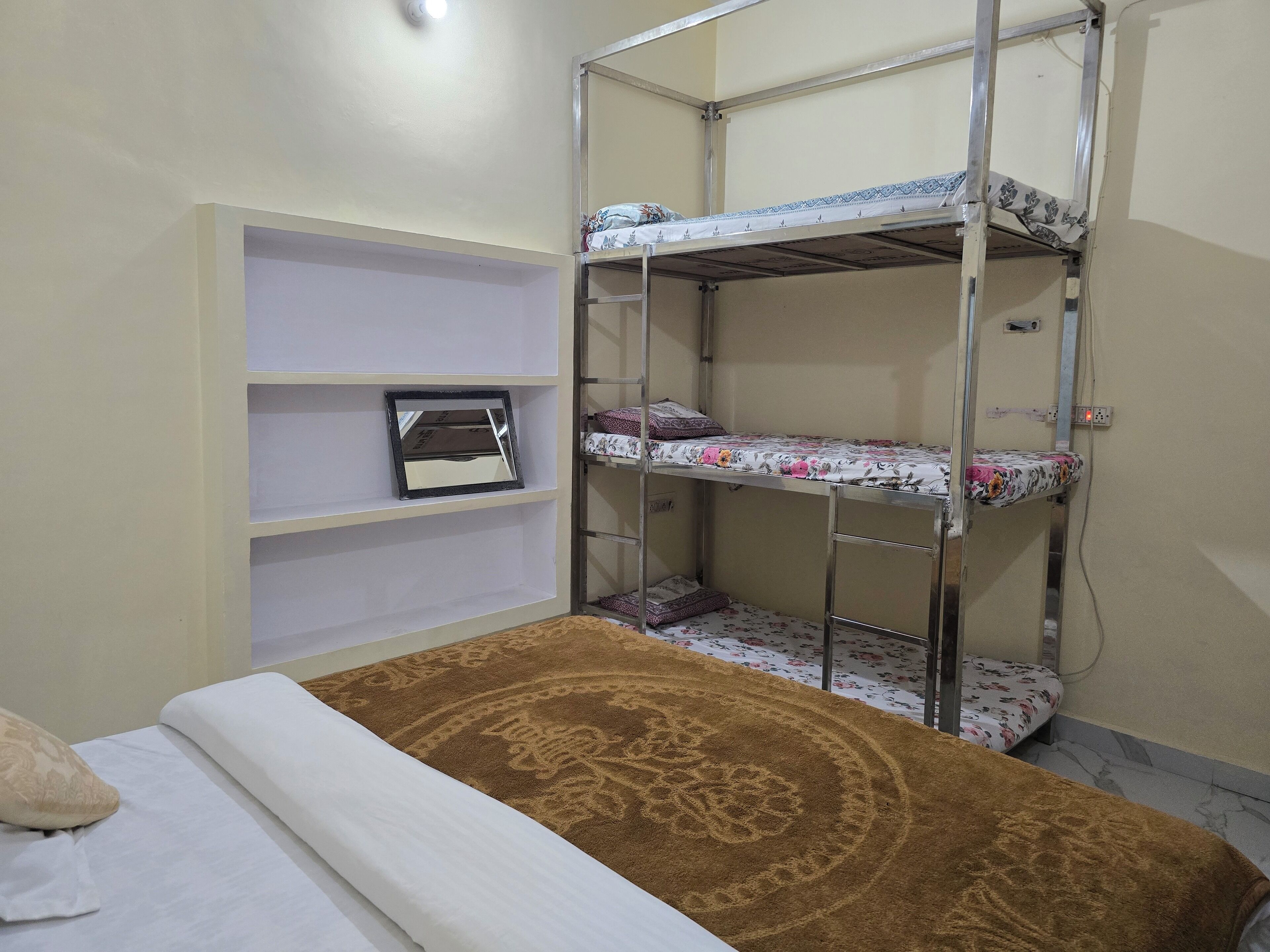Una cama individual con estructura metálica, un colchón blanco y una manta marrón estampada. Se ven dos repisas con mantas dobladas y un cuadro pequeño enmarcados.