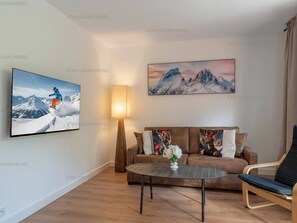 Uma sala de estar com televisão de tela plana, um sofá com almofadas decorativas, uma mesa de centro e um abajur.