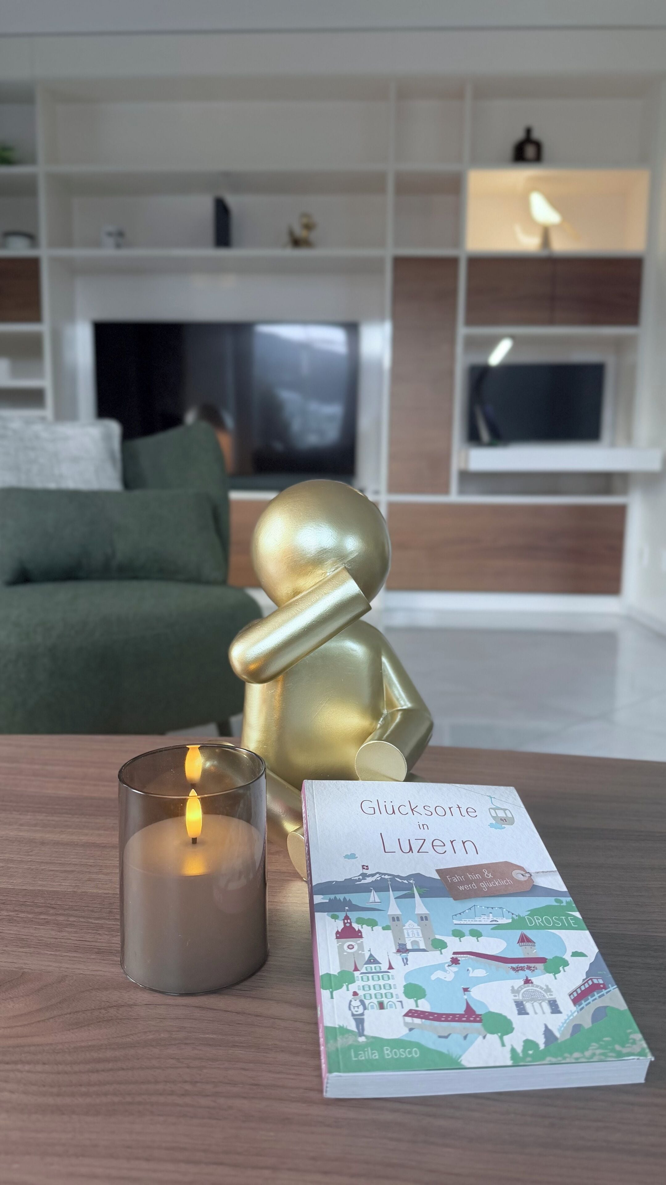 Una sala de estar acogedora con una estatua dorada, una vela encendida y un libro titulado 'Glücksorte in Luzern' de Laila Bosco.